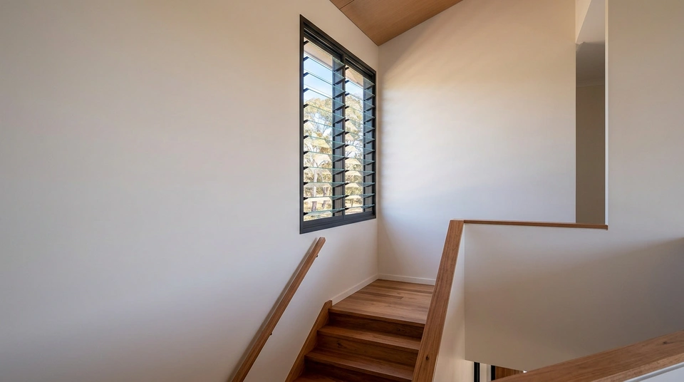 High level aluminium louvre window open above a stair landing in a modern Australian home, with daylight moving air through the tall opening.